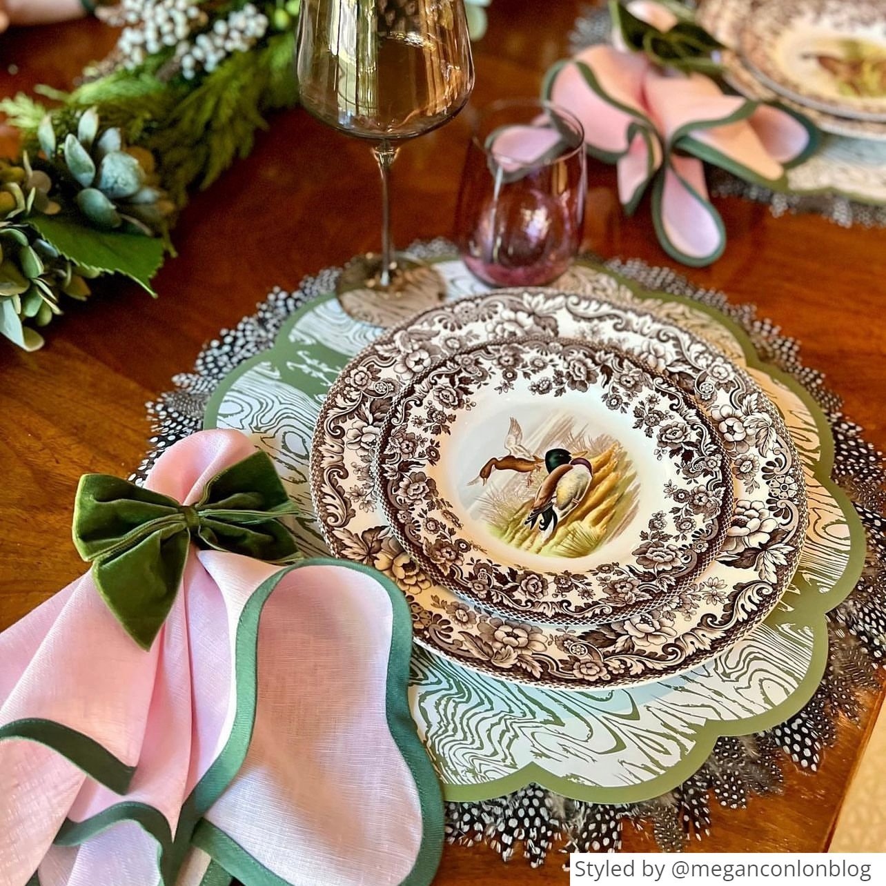 Place setting with a green wood patterned paper placemat layered with duck patterned china on a wooden table with a pink napkin tied with a green felt napkin ring