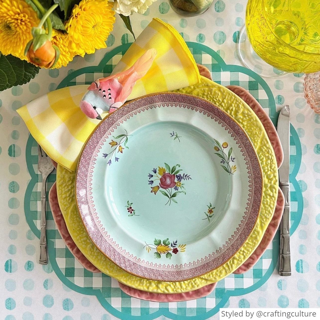 Place setting with green gingham scalloped round paper placemat layered with yellow, pink and white dishes with a yellow napkin tied into a bow