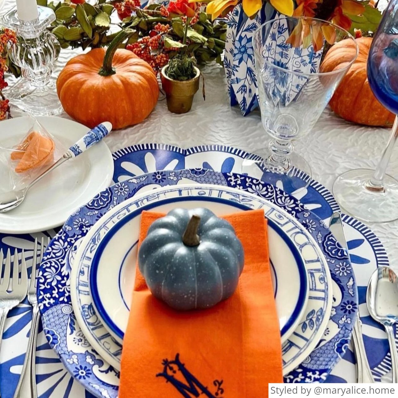 Blue and white scalloped round paper placemat layered with blue and white dishes and an orange napkin with a blue pumpkin on top