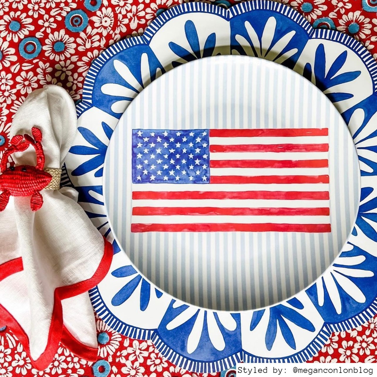 Blue and white scalloped paper placemat layered with an American flag plate and red and white napkin with a crab napkin ring on a red, white and blue floral tablecloth