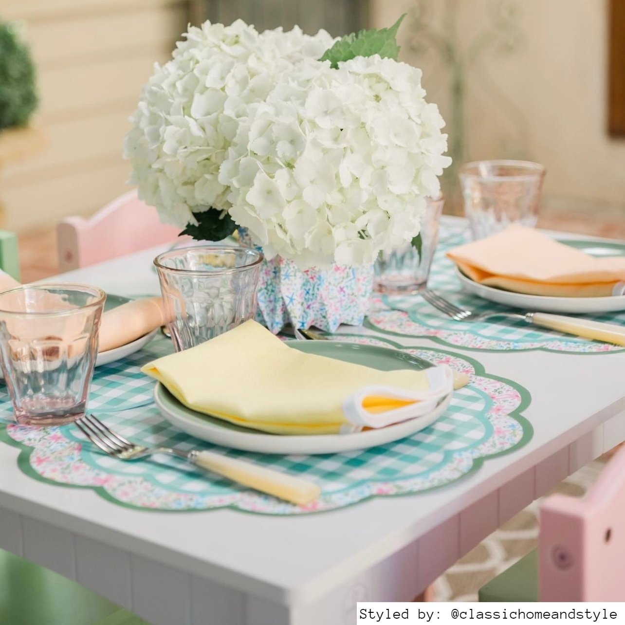 Table setting with multicolored floral scalloped round paper placemats layered with green gingham paper chargers, green plates and a yellow napkin with multicolored floral paper vases and white flowers as centerpieces