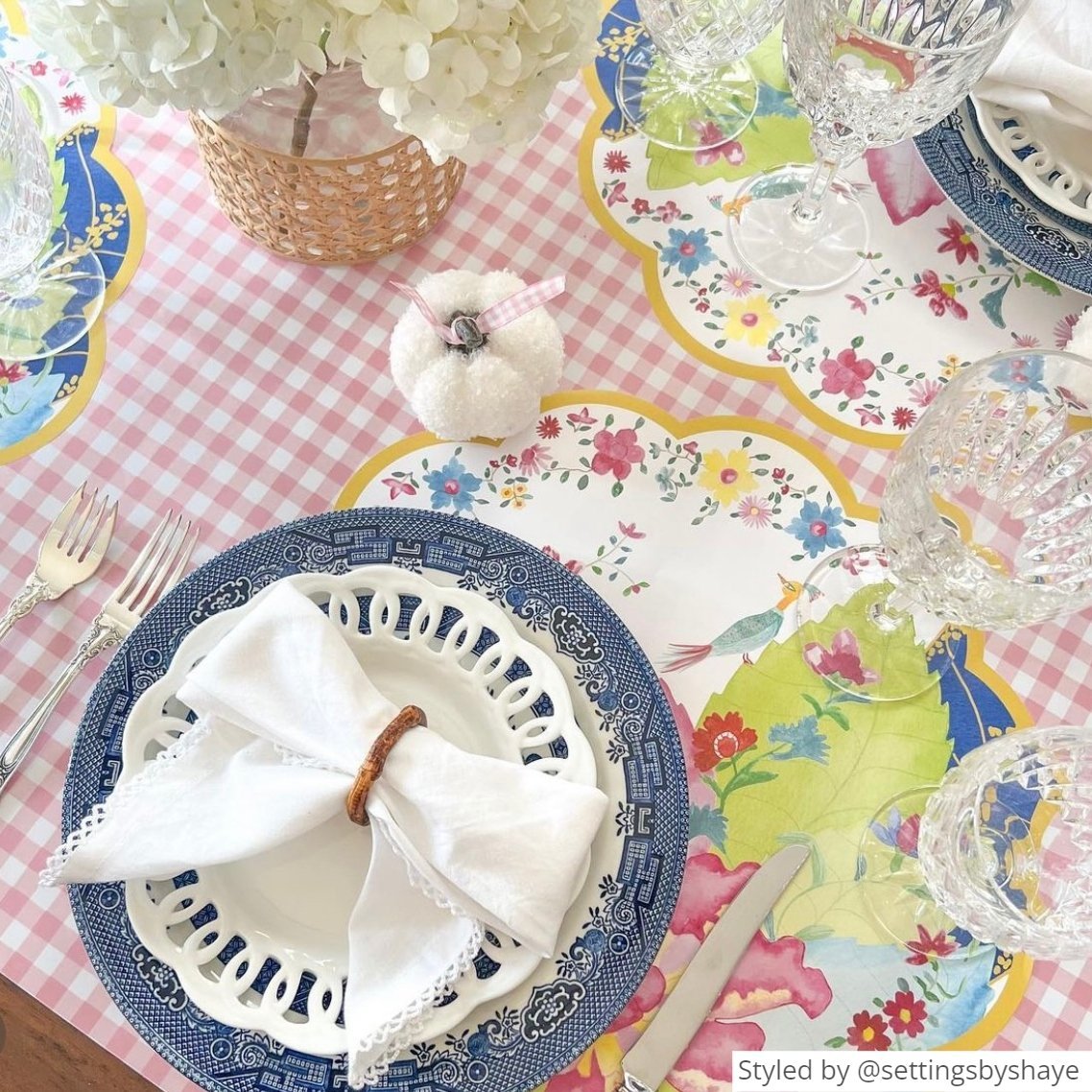 Round scalloped paper placemats featuring a tobacco leaf pattern layered with blue and white plates and a white napkin tied into a bow on top of a pink gingham tablecloth