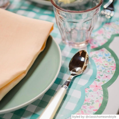 Close up of a place setting with gingham scalloped round paper placemats layered with a multicolored floral scalloped round paper charger and a green plate and yellow napkin