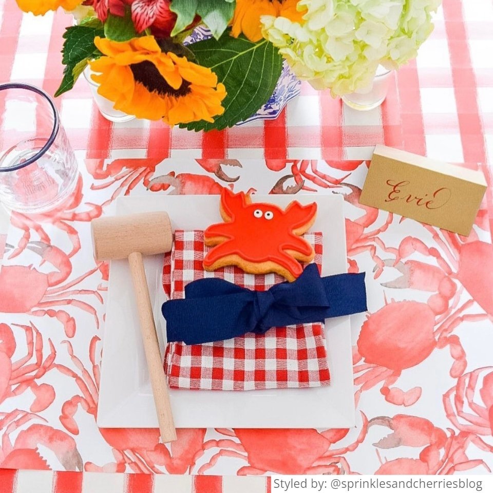 Place setting with a red and white gingham tablecloth layered with crab paper placemats and other crab table decor