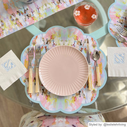 Pink and blue watercolor scalloped round paper placemat layered with a pink plate on a round clear table