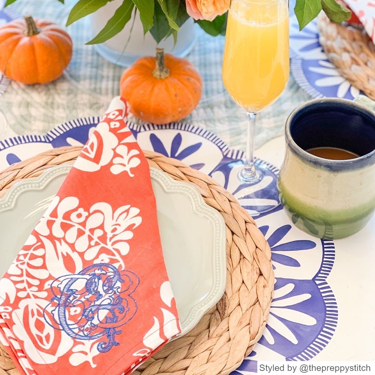 Blue and white scalloped round paper placemat layered with a rattan charger, white plate and orange napkin with mini pumpkins and a mimosa