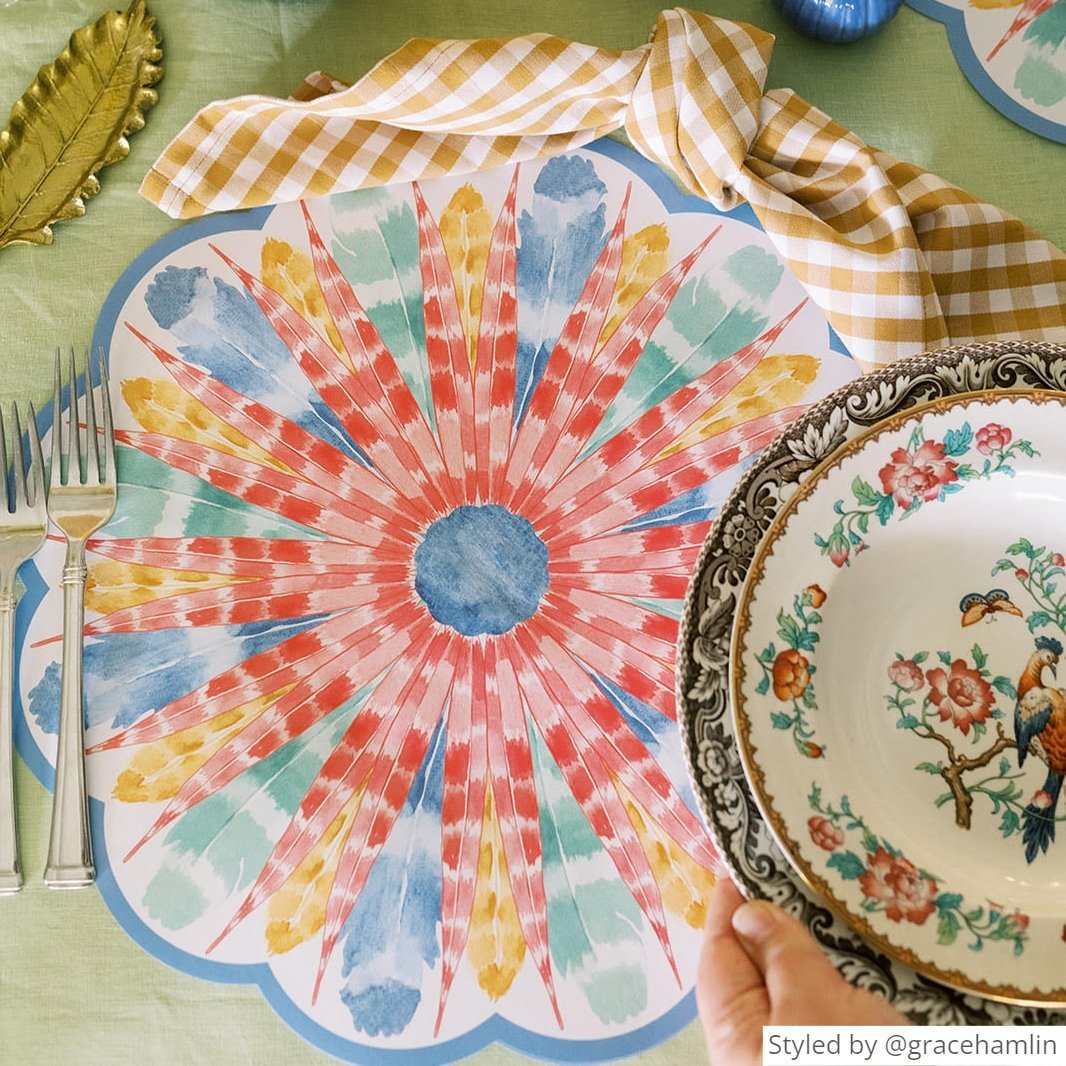 Multicolored feather scalloped paper placemat with a brown gingham napkin on a light green tablecloth with someone placing a stack of plates on top