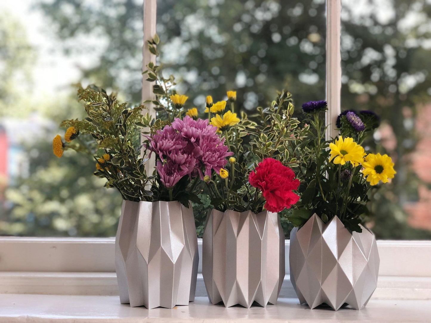 Three silver paper vases with flowers on a windowsill