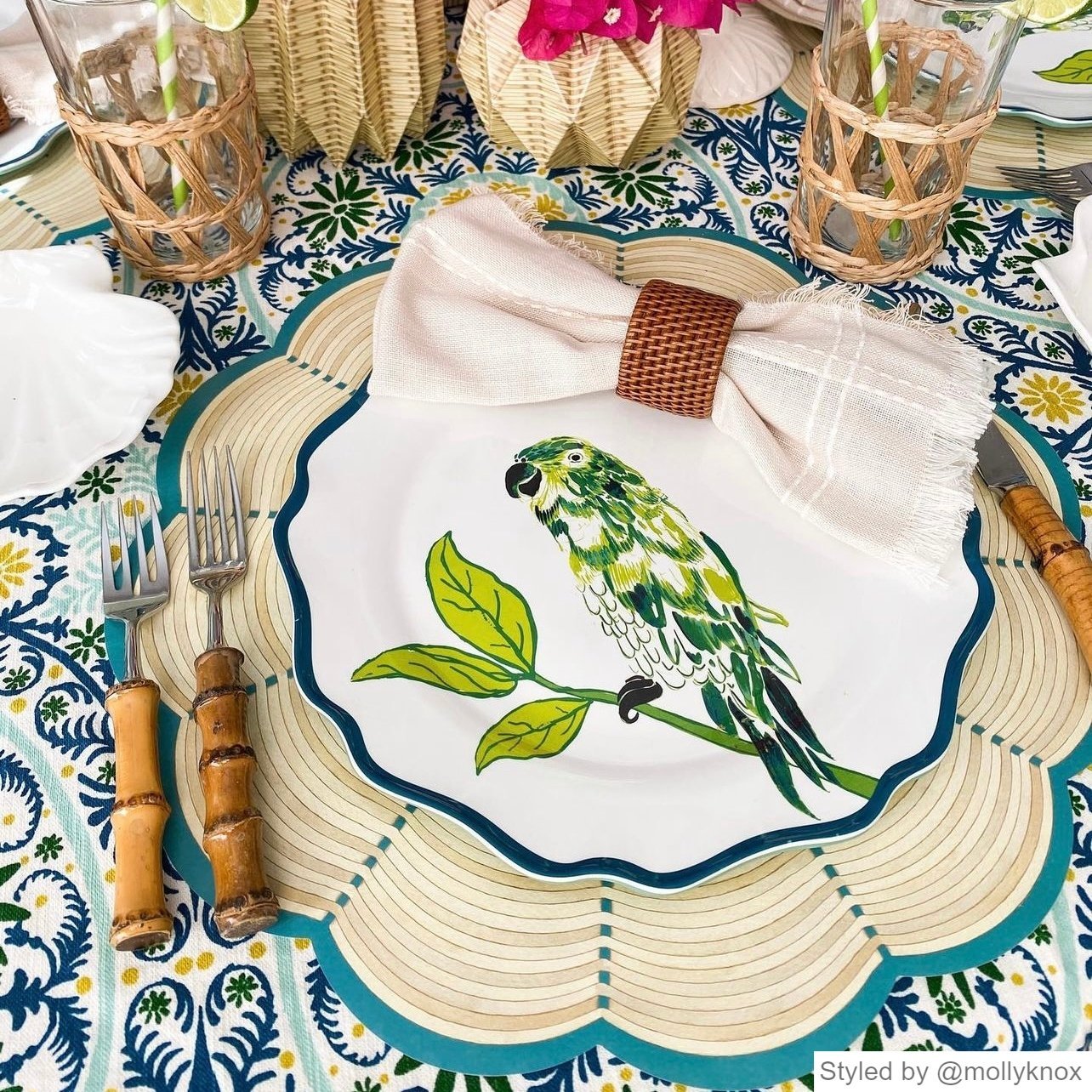 Place setting with a wicker scalloped paper placemat layered with a white plate featuring a parrot, and a white napkin with a rattan napkin ring