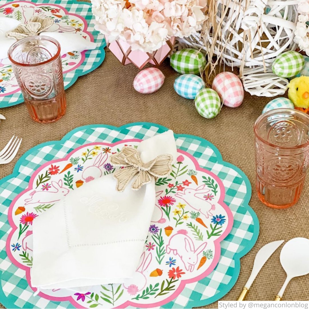 Easter table setting with a green gingham round scalloped paper placemat layered with a bunny paper charger, a white napkin with a rattan bow napkin ring and mini gingham Easter eggs