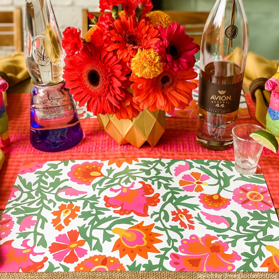 Table setting with a pink, orange and green floral paper placemat on pink tablecloth with gold paper vases and orange flowers