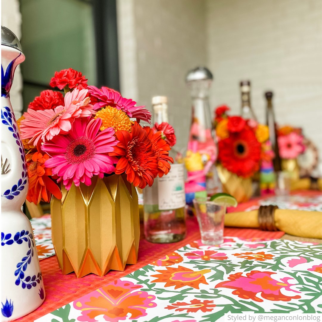 Table setting with a pink, orange and green floral paper placemat on pink tablecloth with gold paper vases and orange flowers