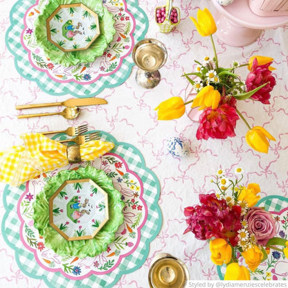 Table setting with green gingham scalloped round paper placemat layered with a bunny paper charger and yellow napkin on a pink tablecloth featuring bows