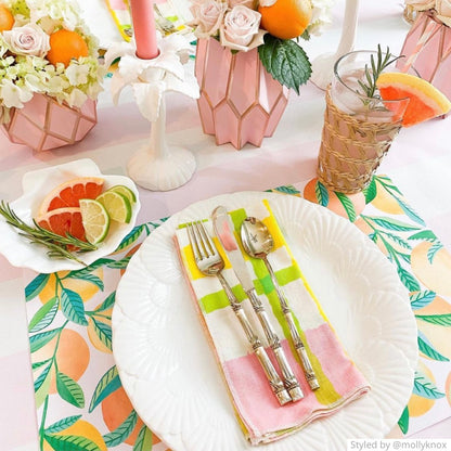 Place setting with a paper placemat featuring oranges on a pink and white tablecloth with fresh fruit and a light pink paper vase centerpiece