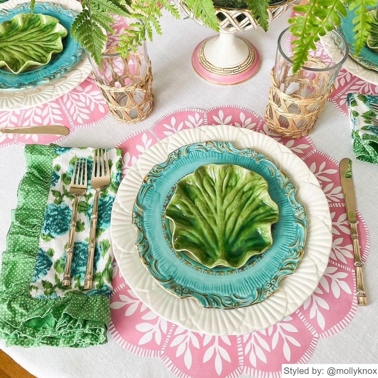Place setting with a pink and white scalloped round paper placemat layered with green, white and blue dishes on a light pink tablecloth with a blue and green napkin