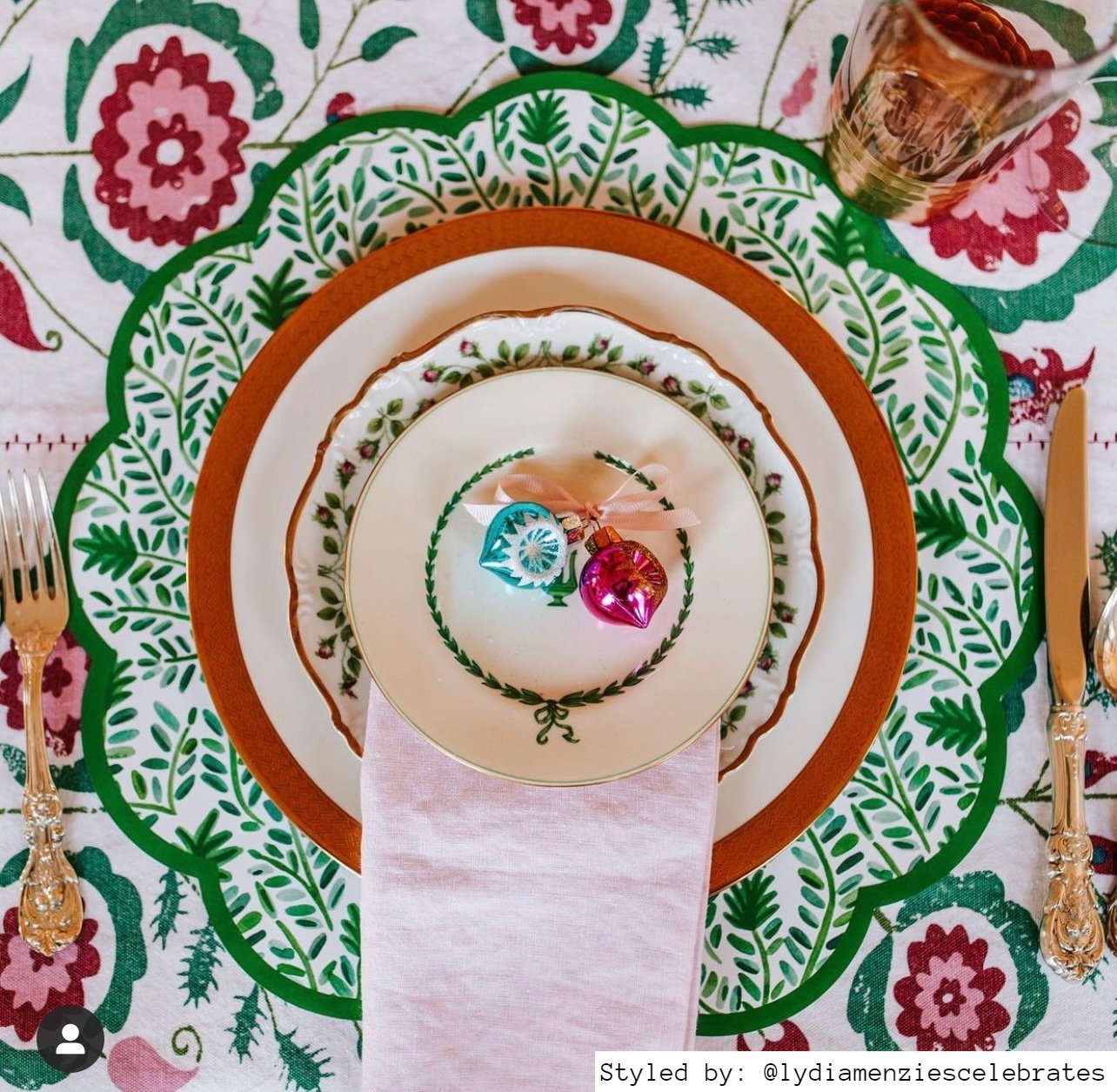 Place setting with a green vine scalloped round paper placemat layered plates and a pink jingle bell on a green and pink floral tablecloth